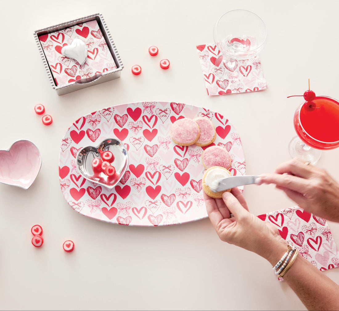 Heart-patterned plate with cookies and a person decorating them on a white surface.