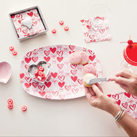 Heart-patterned plate with cookies and a person decorating them on a white surface.